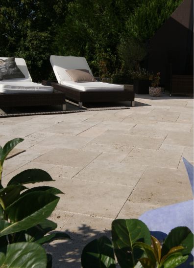 Gemütliche Terrasse mit Travertinplatten verlegt im Römischen Verband mit zwei Liegestühlen und Pflanzen im Vorder- und Hintergrund