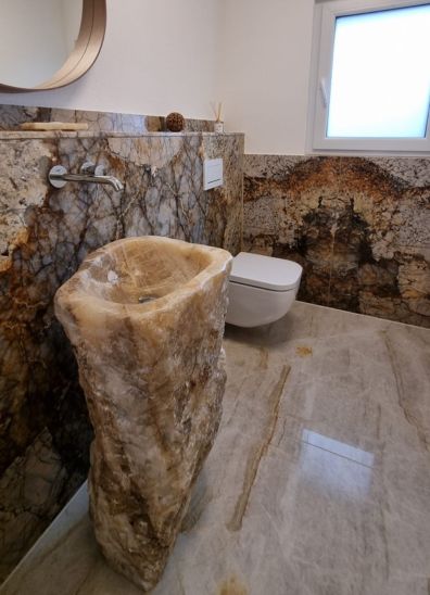 Modernes Badezimmer mit eleganten Natursteinfliesen, die eine luxuriöse und entspannende Atmosphäre schaffen. Ein Standwaschbecken aus Onyx und eine weiße Toilette
