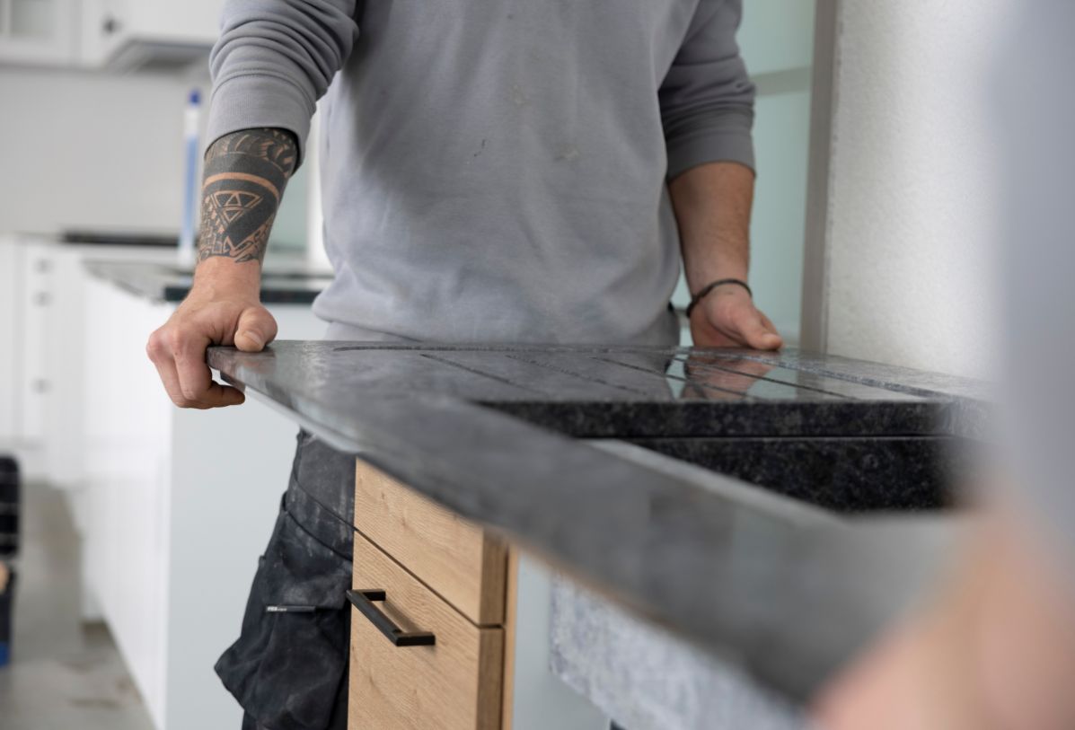 Eine Küchenarbeitsplatte aus einem dunklen Naturstein mit eingeschliffener Abtropffläche wird auf den Küchenkorpus gesetzt.