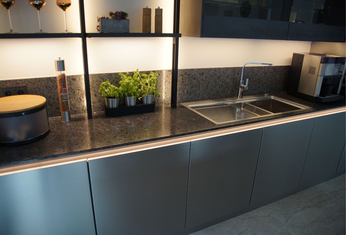 Grauer Küchenkorpus mit einer dunklen Naturstein Küchenarbeitsplatte. Auf der Arbeitsplatte steht eine Kaffeemaschine, ein Spülbecken aus Edelstahl, verschiedene Kräuter, eine Pfeffermühle und ein Brotkorb