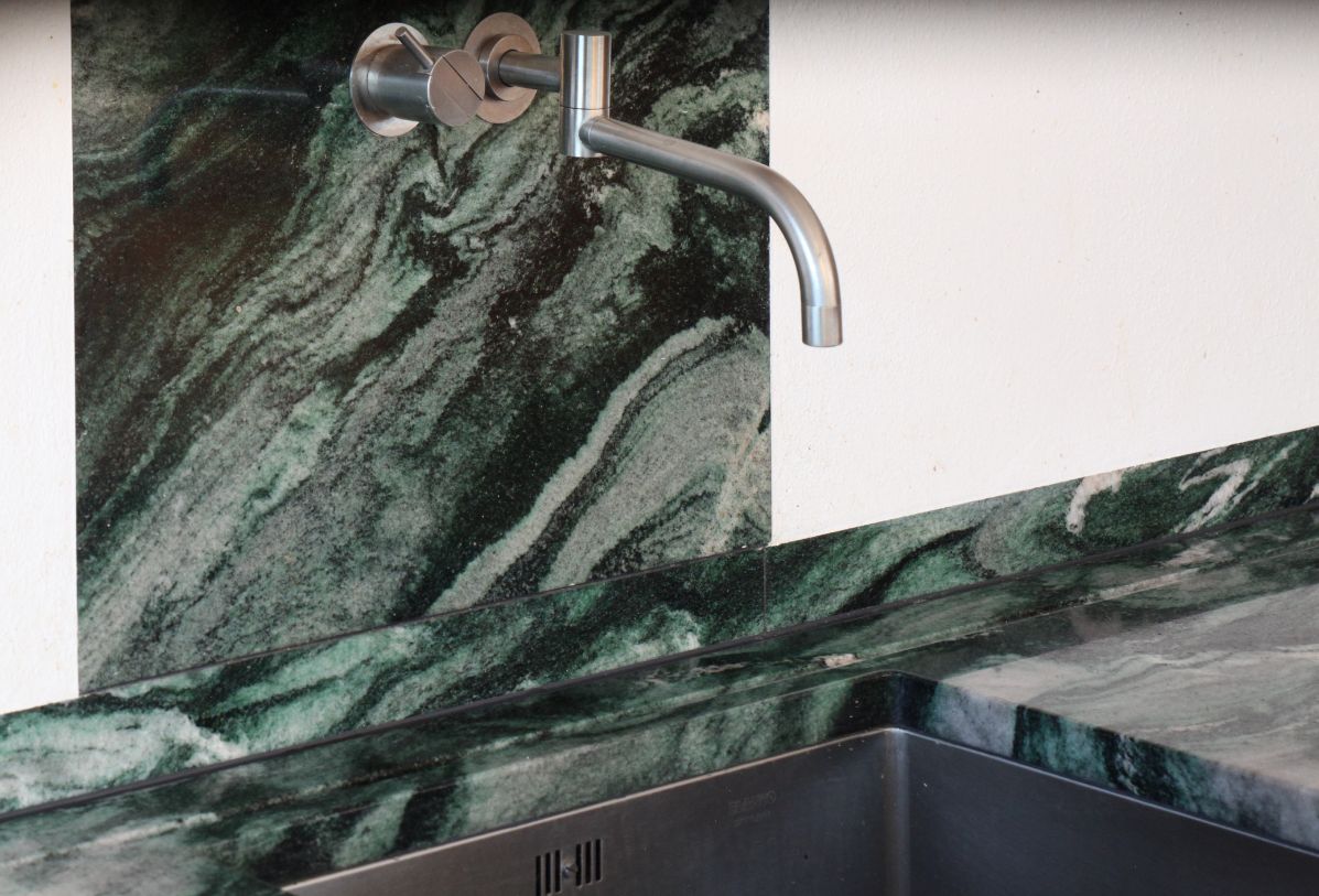 Eine wandhängende Armatur auf einem grünen Naturstein und ein Spülbecken aus Edelstahl als Unterbaubecken in einer Küchenarbeitsplatte aus Naturstein