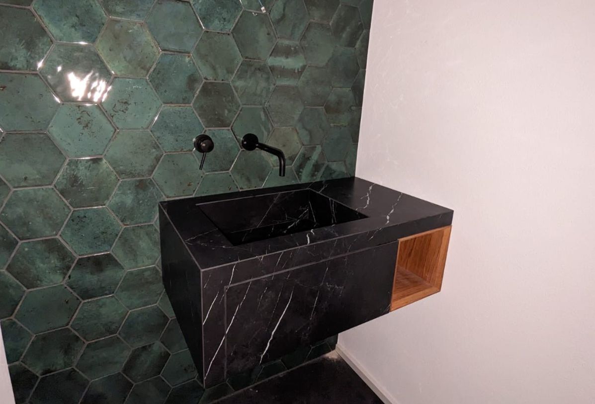 Waschtisch aus dunklem Keramik in Marmoroptik mit eingebautem Ablagefach aus Holz, an der Wand grüne Mosaikplatten und eine schwarze Armatur
